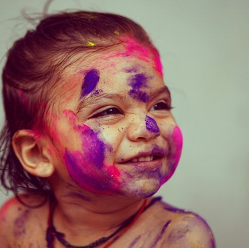 A smiling young child with bright pink and purple paint on their face.