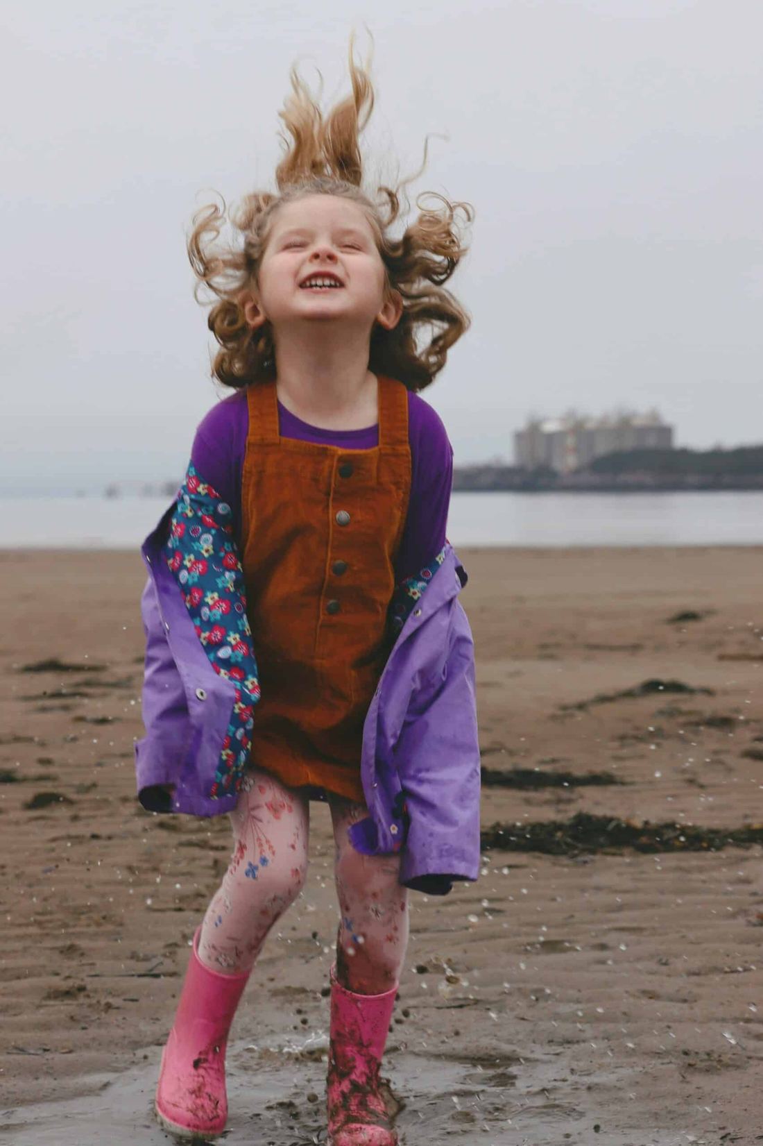 A young girl in a purple raincoat jumping with delight on a beach.