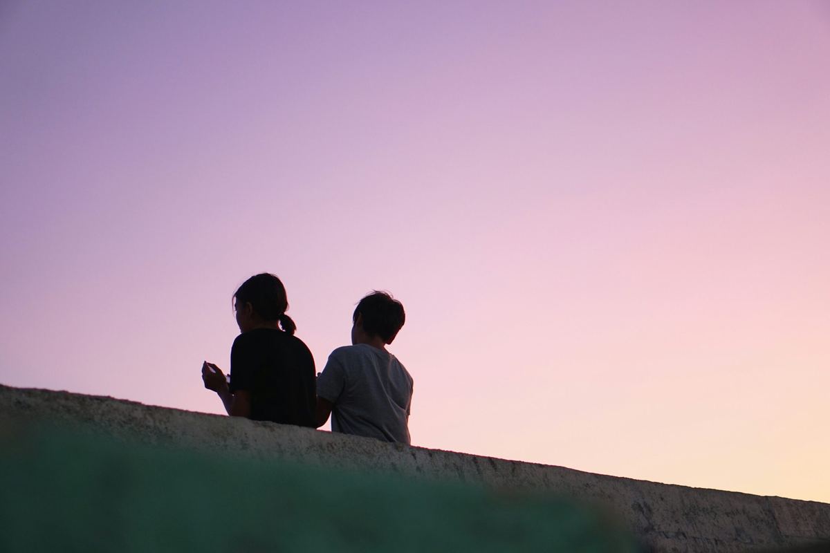 Two young people sitting side by side on a wall against a dusk sky.